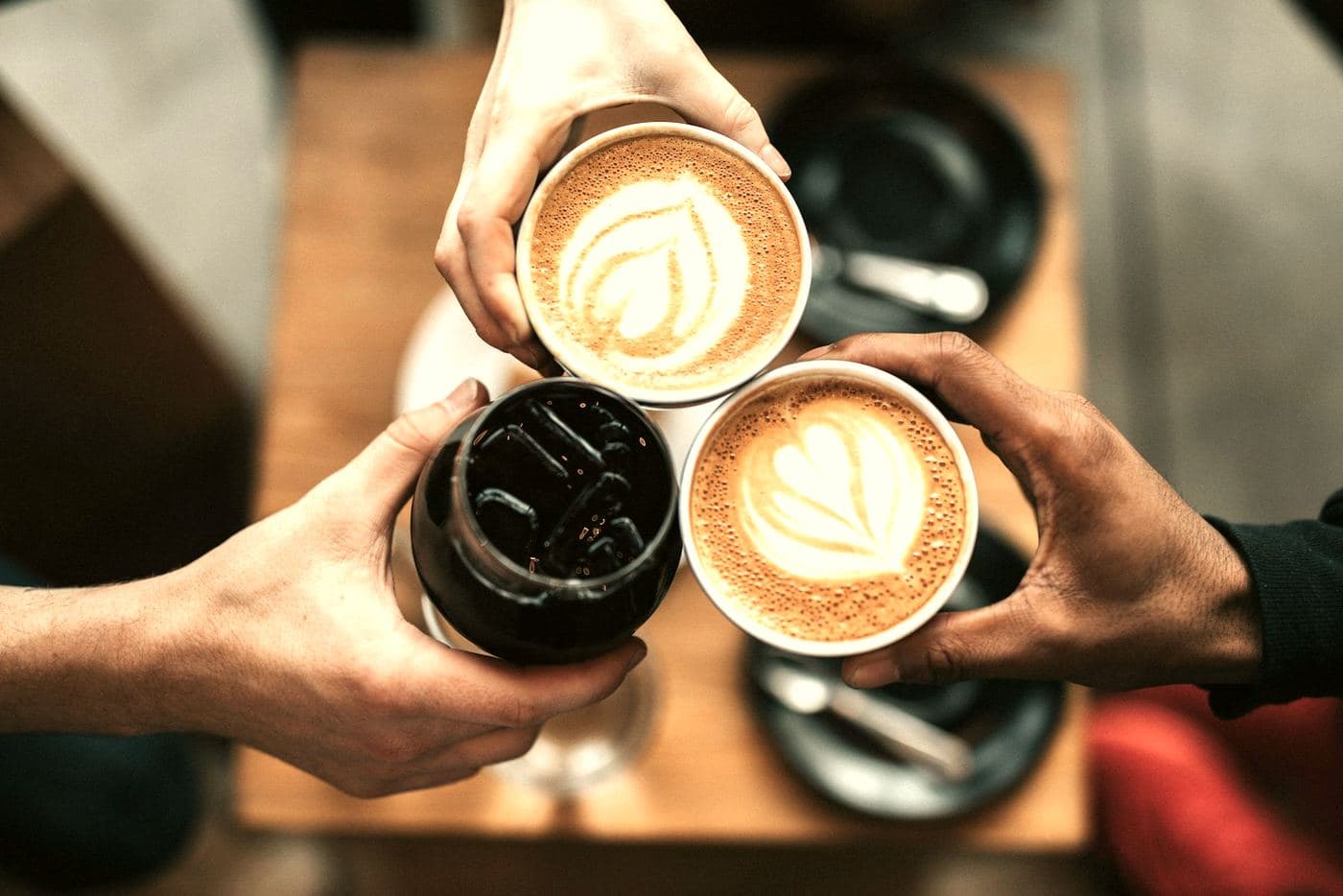 Three hands bringing coffee cups together over a wooden table, seen from above.