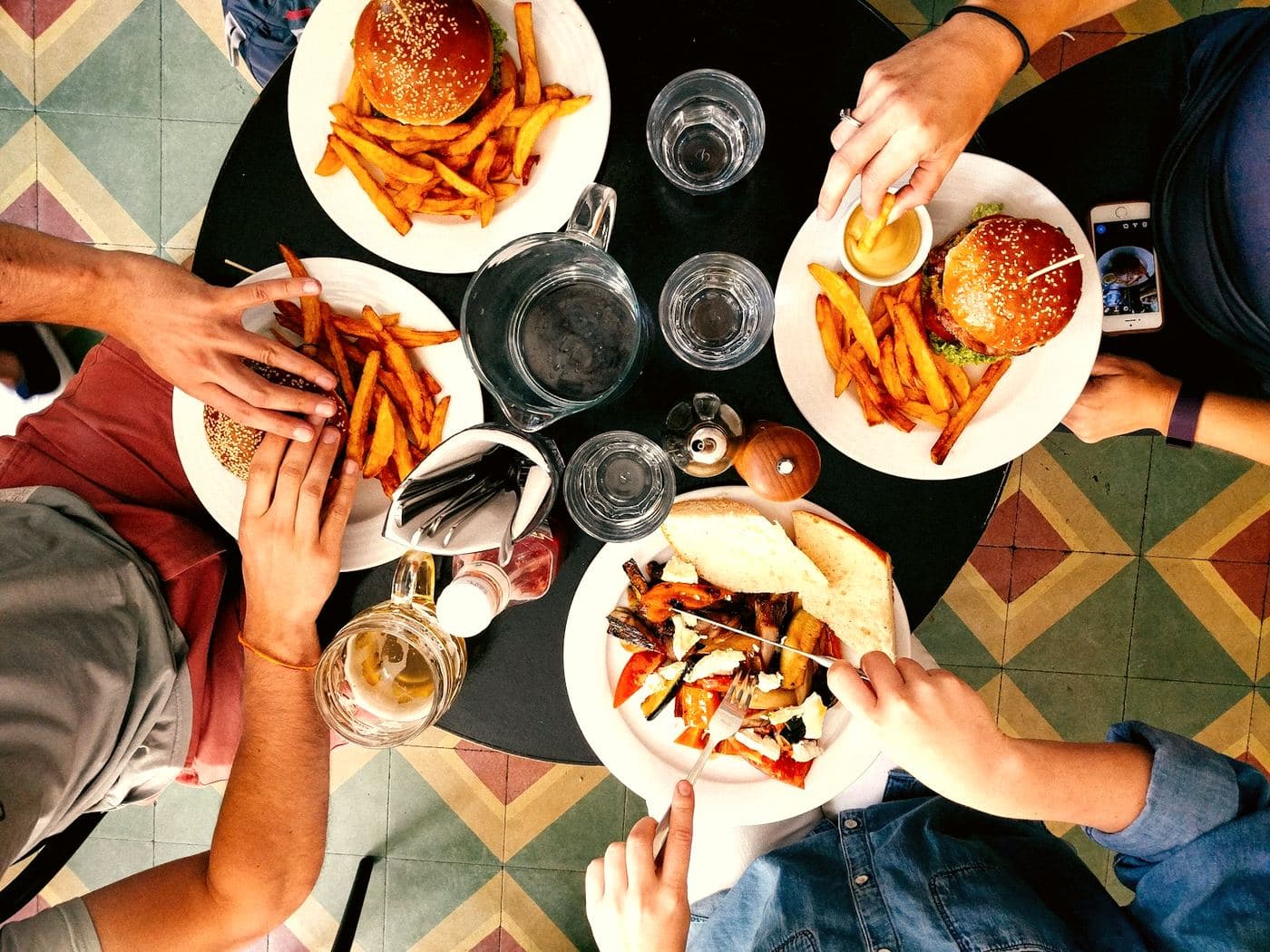 A shared table shot from above, four pairs of hands reaching across plates of burgers and chips.