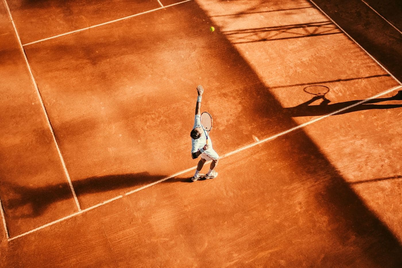 A clay court from above, one player serving and a long shadow across the baseline.